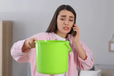 A woman holds a bucket to catch water from a leaky roof while she calls Burroughs Roofing for a repair request